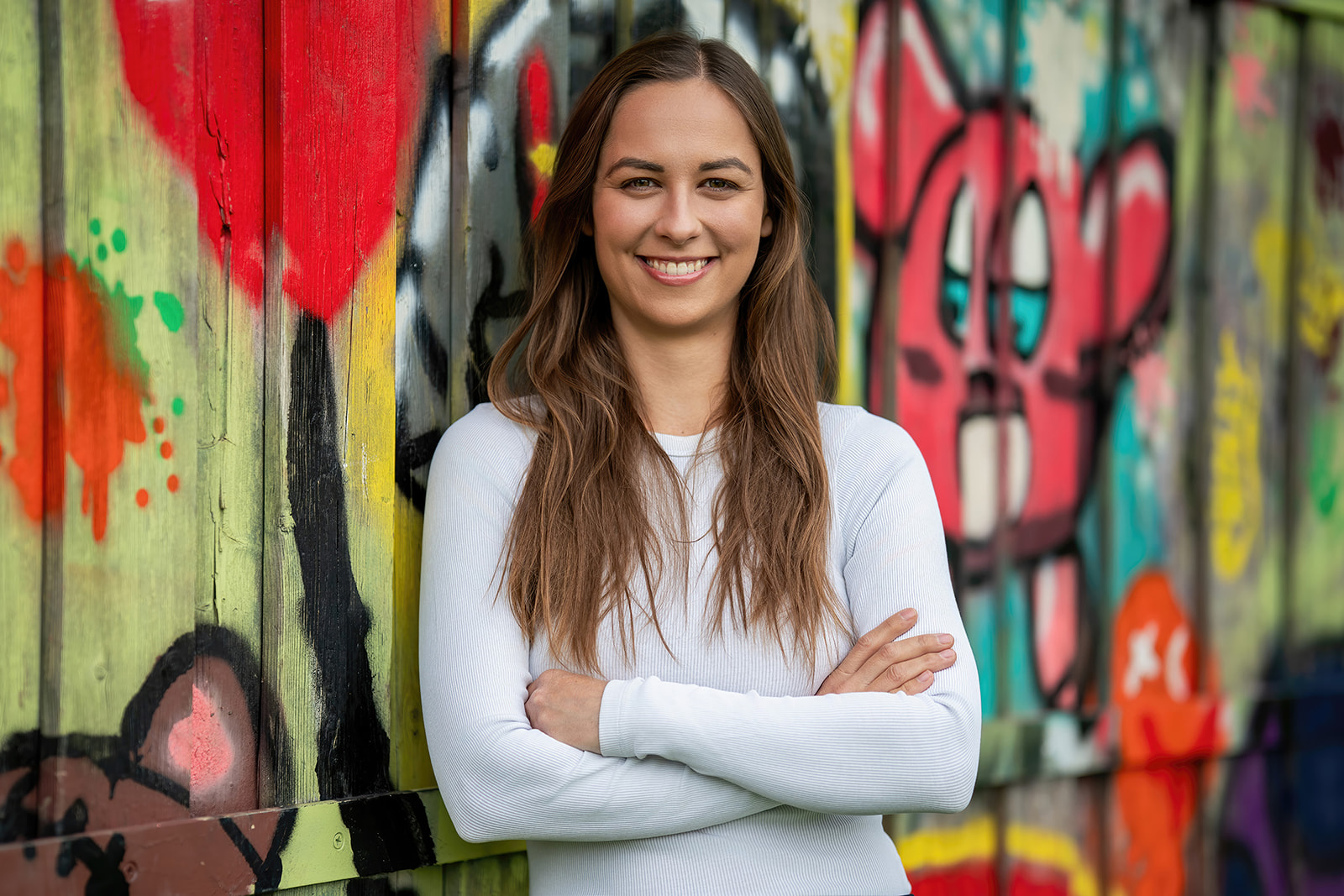 Vor einer farbenfrohen Graffiti-Wand steht die Bürgermeisterkandidatin mit verschränkten Armen – modern, unkonventionell und voller Tatendrang.