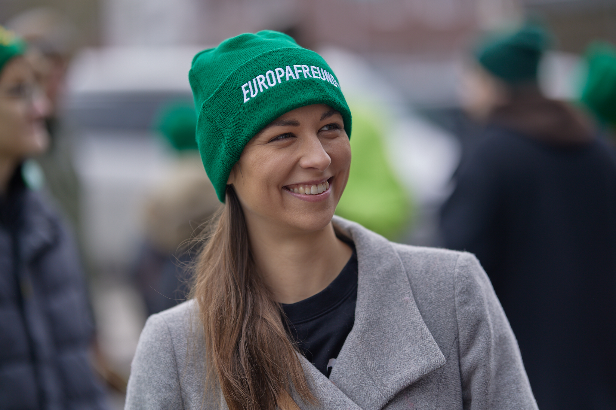 Die Bürgermeisterkandidatin trägt eine grüne Mütze mit der Aufschrift 'EUROPAFREUND*IN' und eine graue Jacke. Sie lächelt und befindet sich inmitten einer Gruppe von Menschen, die ebenfalls grüne Mützen tragen.