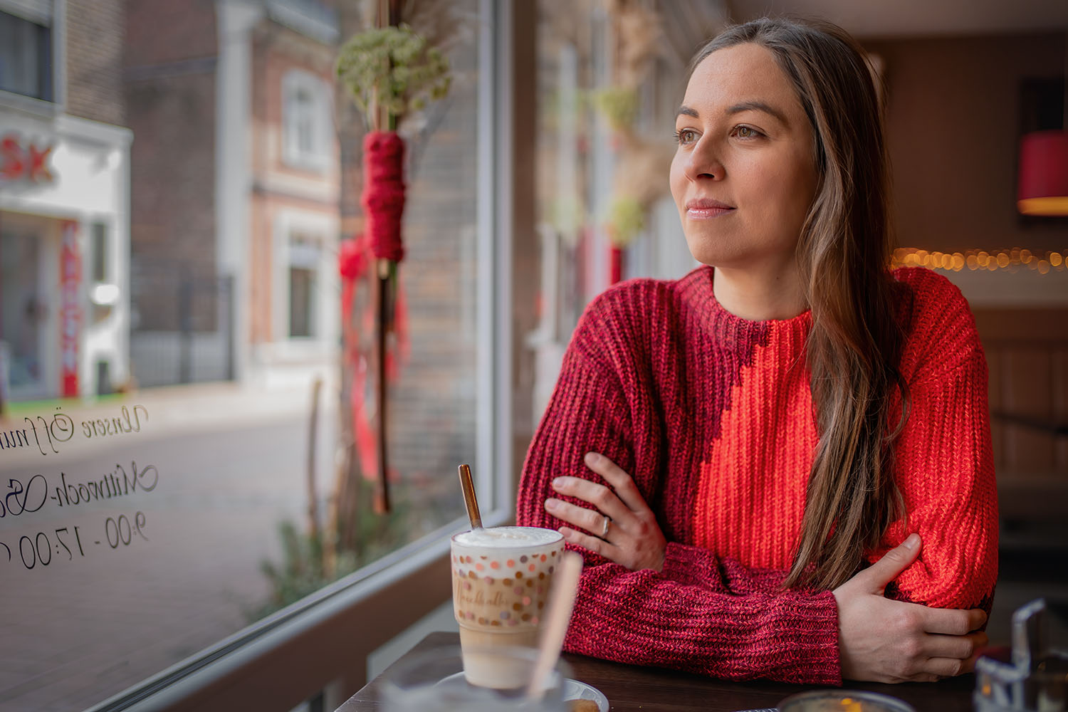 In einem Café, mit nachdenklichem Blick aus dem Fenster, strahlt die Bürgermeisterkandidatin Ruhe und Reflexion aus – ein Moment der Besonnenheit bei einer Tasse Milchkaffee.